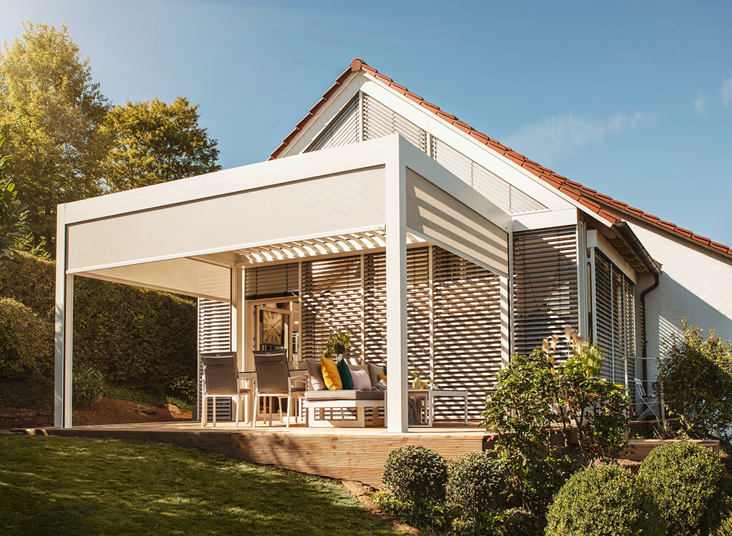 graues-Lamellendach-als-Ueberdachung-fuer-eine-Aussen-Terrasse-mit-Sitzmoeglichkeiten-an-einem-weissen-Haus